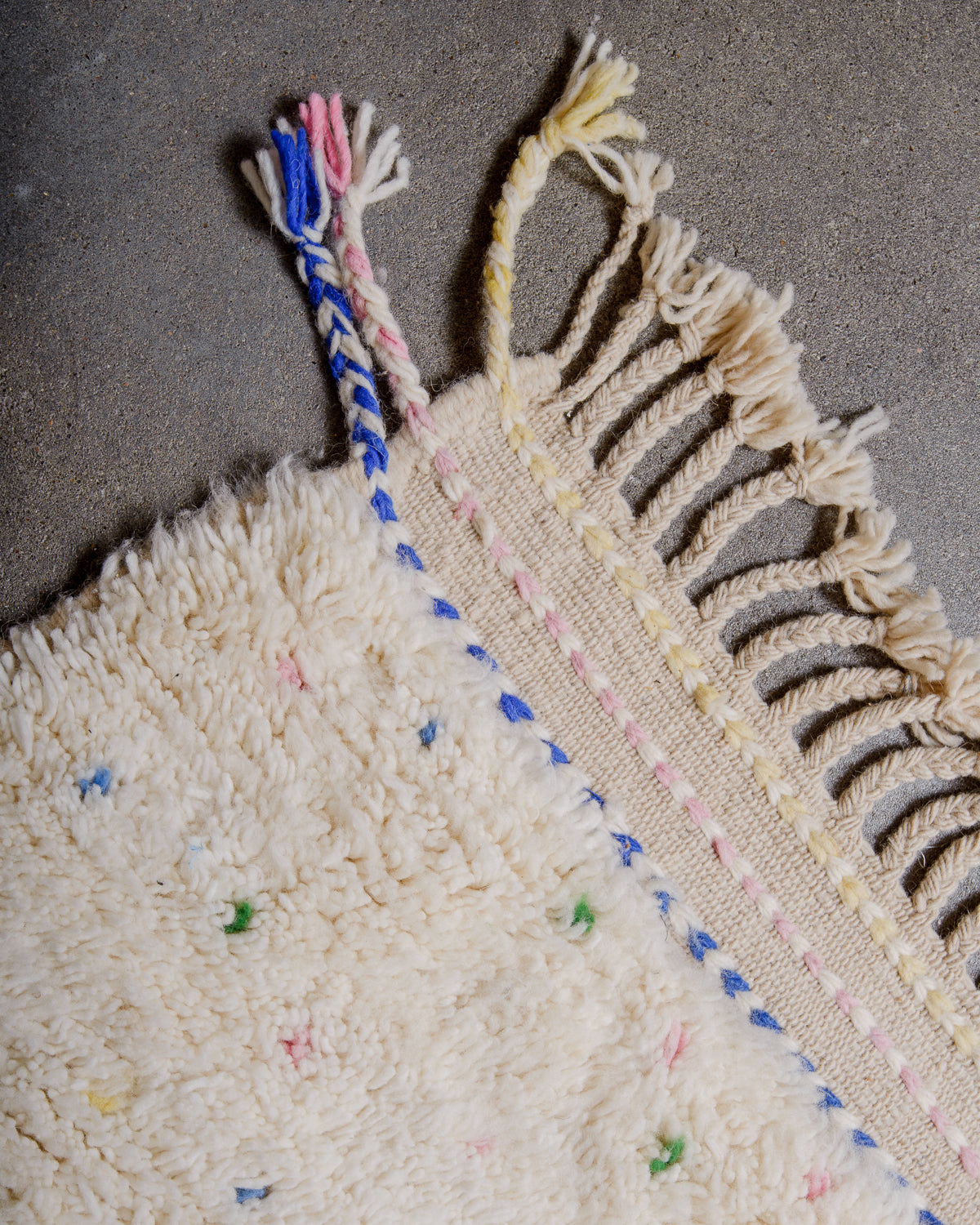 Corner of Berber rug with blue, pink and yellow tassels at the corner and natural wool tassels across the top.
